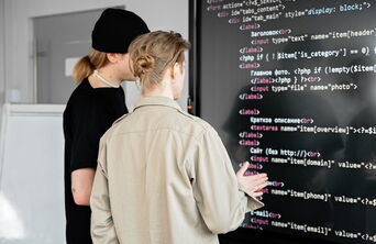 Two professional web developers from the internet agency 5150media review and write clean HTML5 source code on a large monitor to optimize a client's website.